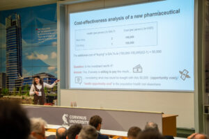A presenter gestures toward a slide titled "Cost-effectiveness analysis of a new pharmaceutical" at Corvinus University.